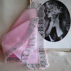 May include: A pink handkerchief with a delicate silver lace trim. The handkerchief is folded and displayed on a table.