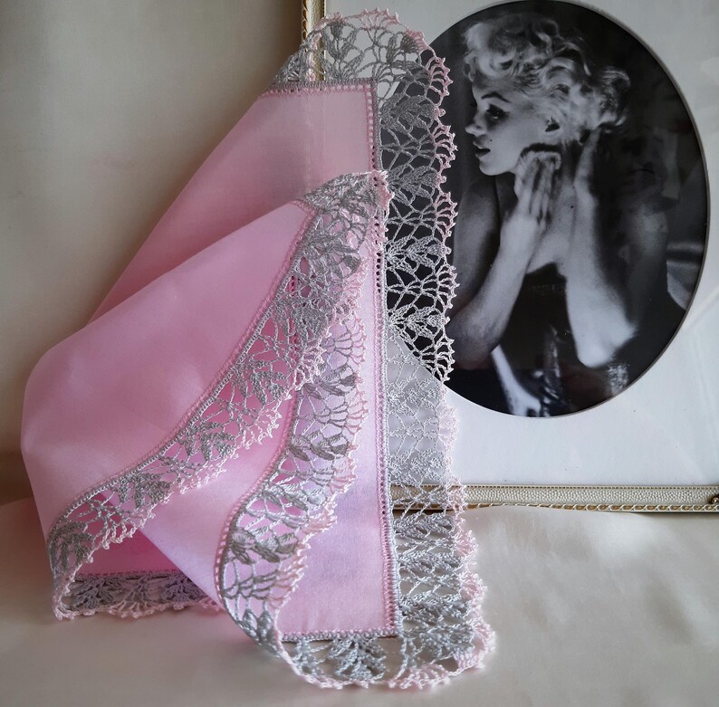May include: A pink handkerchief with a delicate silver lace trim. The handkerchief is folded and displayed on a table.