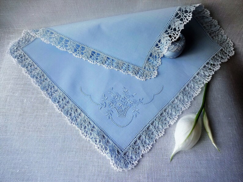 May include: A light blue handkerchief with white crochet trim and an embroidered floral design. The handkerchief is folded and lying on a light blue textured surface.