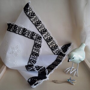 May include: A white handkerchief with black crocheted trim and a white embroidered floral design. A white porcelain bird with a long tail feather and a gold safety pin with a blue gemstone are also in the image.