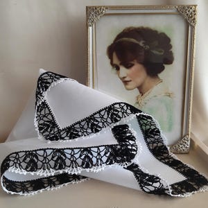 May include: A white handkerchief with a black crocheted lace border. The handkerchief is folded and lying on a white surface.