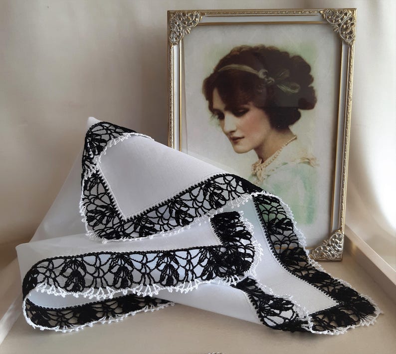 May include: A white handkerchief with a black crocheted lace border. The handkerchief is folded and lying on a white surface.