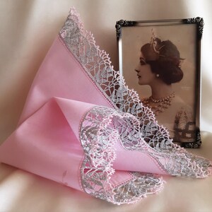 May include: A pink handkerchief with a silver lace trim. The handkerchief is folded in a triangle shape and is lying on a white surface.