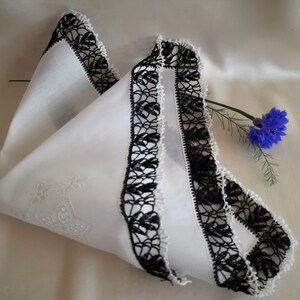 May include: White cotton handkerchief with black crocheted lace trim. The handkerchief is folded in half and has a floral design embroidered in the center.