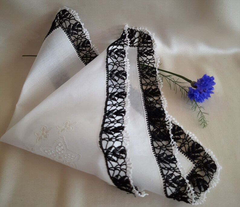 May include: White cotton handkerchief with black crocheted lace trim. The handkerchief is folded in half and has a floral design embroidered in the center.