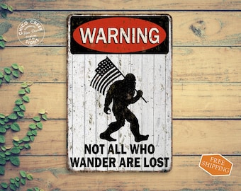 May include: A white metal sign with a red warning sign at the top. The sign features a black silhouette of Bigfoot carrying an American flag and the text "WARNING NOT ALL WHO WANDER ARE LOST".