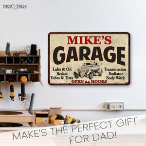 May include: A vintage-style metal sign with the text "MIKE'S GARAGE" in black lettering on a cream background. The sign features a black and white illustration of a classic car and the text "Lube & Oil, Brakes, Tubes & Tires, Transmission, Radiator, Body Work, OPEN 24 HOURS" in red lettering.