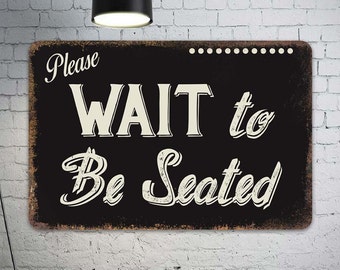 May include: Black and white metal sign with a distressed look. The sign reads "Please WAIT to Be Seated".