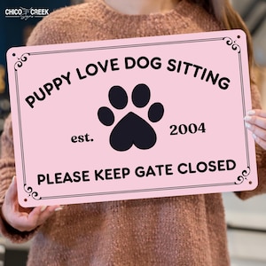 May include: A pink metal sign with black text that reads "PUPPY LOVE DOG SITTING" above a paw print and the year "2004". The bottom of the sign says "PLEASE KEEP GATE CLOSED". Decorative swirls are in each corner.