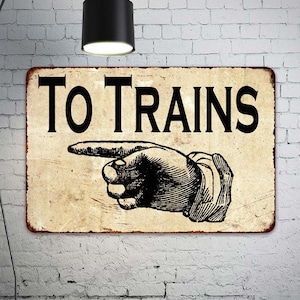 May include: A vintage-style metal sign with a distressed, aged look. The sign reads "TO TRAINS" in black lettering and features a black and white illustration of a pointing hand.