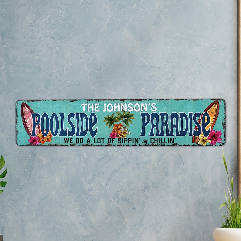Pode incluir: Uma placa azul turquesa com o texto "The Johnson's Poolside Paradise We do a lot of sippin' & chillin'" em branco. A placa apresenta duas pranchas de surf e flores tropicais.