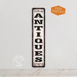 May include: A vintage-style metal sign with a distressed white background and black lettering that reads "ANTIQUES". The sign has a rustic, weathered look and is perfect for adding a touch of charm to any home or business.