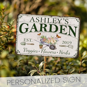 Peut inclure: Une pancarte de jardin personnalis&eacute;e avec un fond blanc et du texte vert. La pancarte indique "Ashley's Garden Est. 2019 Veggies. Flowers. Herbs". La pancarte pr&eacute;sente &eacute;galement une brouette avec des fleurs et des papillons.