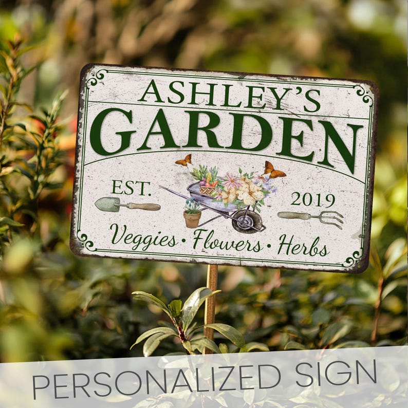 Peut inclure: Une pancarte de jardin personnalis&eacute;e avec un fond blanc et du texte vert. La pancarte indique "Ashley's Garden Est. 2019 Veggies. Flowers. Herbs". La pancarte pr&eacute;sente &eacute;galement une brouette avec des fleurs et des papillons.