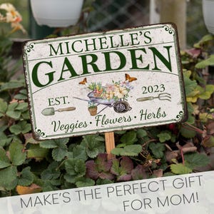 Peut inclure: Une pancarte de jardin en m&eacute;tal blanc avec du texte vert qui dit "Michelle's Garden Est. 2023 Veggies Flowers Herbs". La pancarte a un aspect rustique et vieilli et pr&eacute;sente une petite brouette avec des fleurs et des papillons.