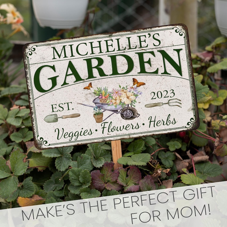 Peut inclure: Une pancarte de jardin en m&eacute;tal blanc avec du texte vert qui dit "Michelle's Garden Est. 2023 Veggies Flowers Herbs". La pancarte a un aspect rustique et vieilli et pr&eacute;sente une petite brouette avec des fleurs et des papillons.