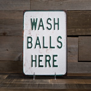 Peut inclure: Un panneau en m&eacute;tal blanc avec du texte vert qui dit "WASH BALLS HERE" sur un fond en bois rustique.