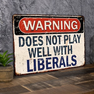 May include: A vintage-style metal sign with a warning sign design. The sign is white with red and blue text. The text reads "WARNING DOES NOT PLAY WELL WITH LIBERALS".