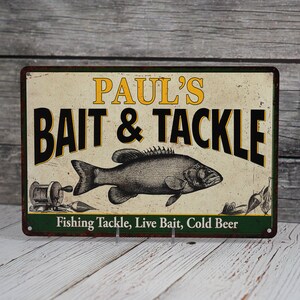 May include: A vintage-style metal sign with a green border and a white background. The sign reads "Paul's Bait & Tackle" in black text with a yellow outline. A black and white illustration of a fish is in the center of the sign. The text "Fishing Tackle, Live Bait, Cold Beer" is printed in black at the bottom of the sign.