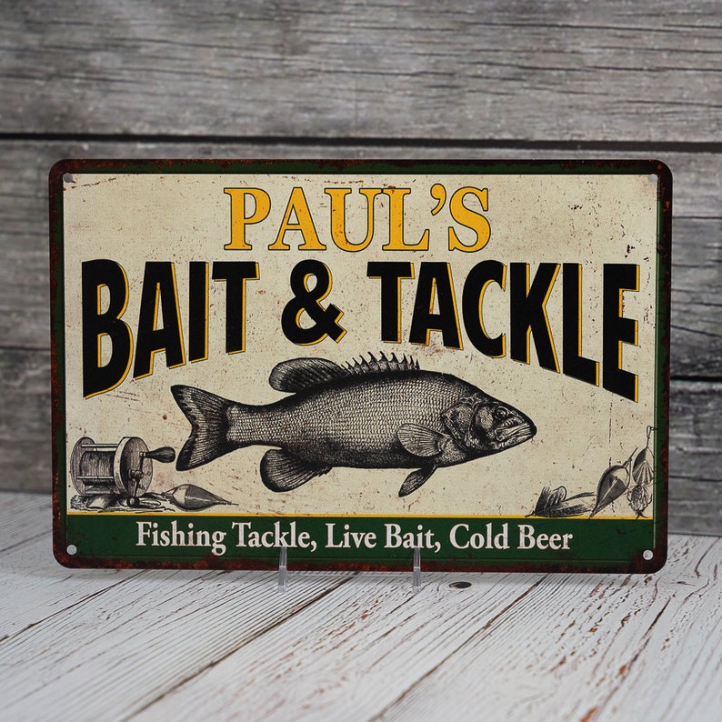 May include: A vintage-style metal sign with a green border and a white background. The sign reads "Paul's Bait & Tackle" in black text with a yellow outline. A black and white illustration of a fish is in the center of the sign. The text "Fishing Tackle, Live Bait, Cold Beer" is printed in black at the bottom of the sign.