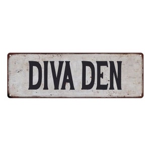 May include: A white metal sign with a rusty brown border. The sign reads "Diva Den" in black lettering.