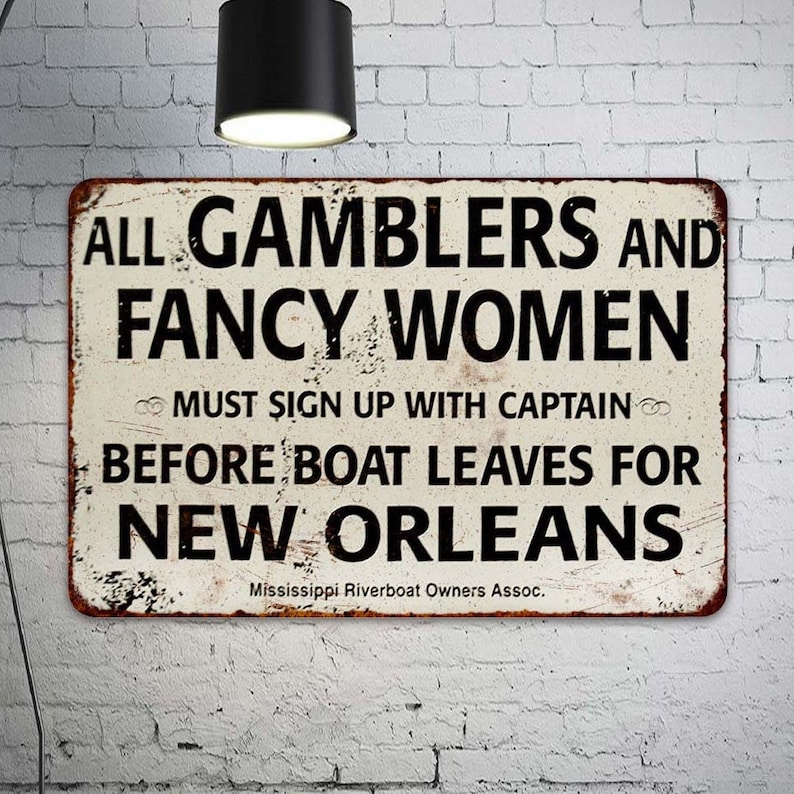 May include: A vintage-style metal sign with a white background and black text. The sign reads "ALL GAMBLERS AND FANCY WOMEN MUST SIGN UP WITH CAPTAIN BEFORE BOAT LEAVES FOR NEW ORLEANS" and "Mississippi Riverboat Owners Assoc."