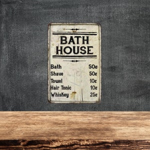May include: A vintage-style metal sign with the text "BATH HOUSE" and a price list for bath, shave, towel, hair tonic, and whiskey. The sign is white with black lettering and is slightly worn, giving it a rustic look.