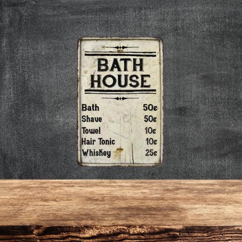 May include: A vintage-style metal sign with the text "BATH HOUSE" and a price list for bath, shave, towel, hair tonic, and whiskey. The sign is white with black lettering and is slightly worn, giving it a rustic look.