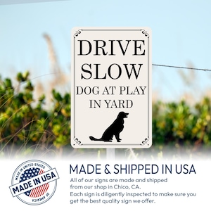 May include: A rectangular sign with the text "DRIVE SLOW DOG AT PLAY IN YARD" in black. A black silhouette of a dog is at the bottom. The sign has decorative flourishes and is set against a blurred outdoor background.
