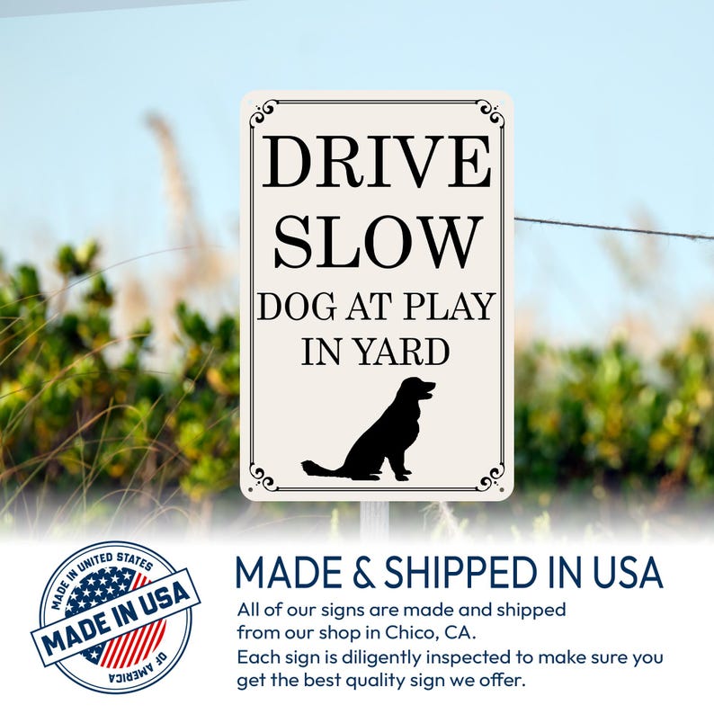 May include: A rectangular sign with the text "DRIVE SLOW DOG AT PLAY IN YARD" in black. A black silhouette of a dog is at the bottom. The sign has decorative flourishes and is set against a blurred outdoor background.