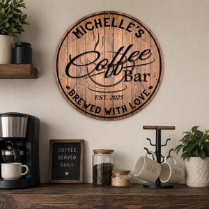 May include: A round wooden sign with the text "MICHELLE'S Coffee Bar" and "BREWED WITH LOVE" in black lettering. The sign also includes "EST. 2025". The sign is hanging above a coffee station with a coffee maker, mugs, and coffee beans.