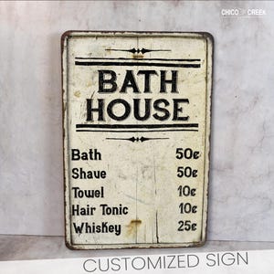 May include: A vintage-style metal sign with a white background and black text that reads "Bath House". The sign lists prices for various services, including bath, shave, towel, hair tonic, and whiskey.