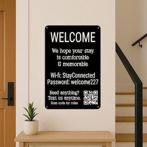 Op de afbeelding: Zwart rechthoekig bord met witte tekst "WELCOME". Het bord bevat Wi-Fi-informatie en een QR-code. Het is aan een muur bevestigd boven een houten tafel.