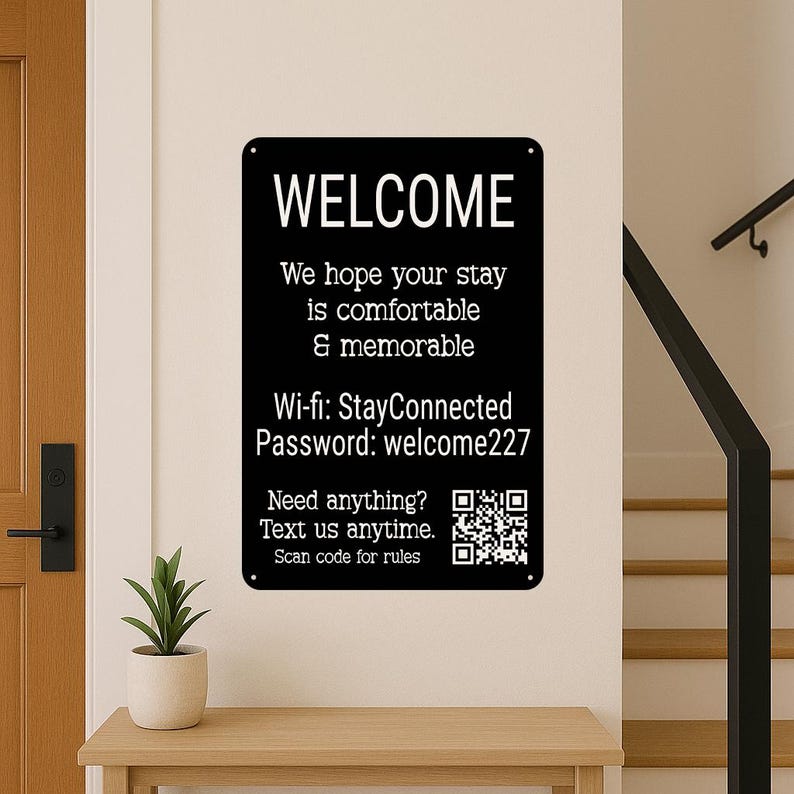 Op de afbeelding: Zwart rechthoekig bord met witte tekst "WELCOME". Het bord bevat Wi-Fi-informatie en een QR-code. Het is aan een muur bevestigd boven een houten tafel.