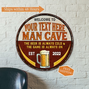 May include: A round, rusty metal sign with a brown background and yellow accents. The sign says "Welcome to Your Text Here Man Cave" with a beer mug graphic and the text "The beer is always cold & the game is always on. Est 2022."