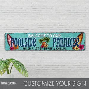 Pode incluir: Uma placa de metal azul turquesa com o texto "Welcome to our Poolside Paradise We do a lot of sippin' & chillin'" em branco. A placa apresenta duas pranchas de surf com flores tropicais e palmeiras.