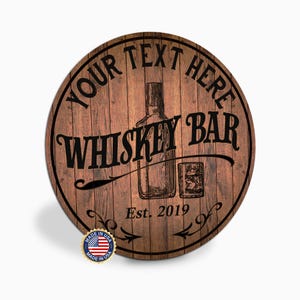 May include: Round wooden sign with a whiskey bar theme. The sign features the text "YOUR TEXT HERE" at the top, "WHISKEY BAR" in the center, and "Est. 2019" at the bottom. A whiskey bottle and glass are illustrated in the center. Includes a "Made in USA" sticker.