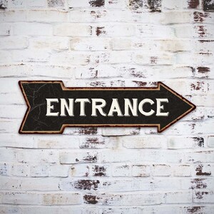 May include: A black and brown metal arrow sign with the word "ENTRANCE" in white letters on a white brick wall background.