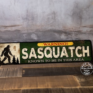 May include: A rectangular metal sign with a distressed finish. The sign features a silhouette of Bigfoot, the text "WARNING! SASQUATCH KNOWN TO BE IN THIS AREA," and a logo. The color scheme includes green, yellow, white, and brown.