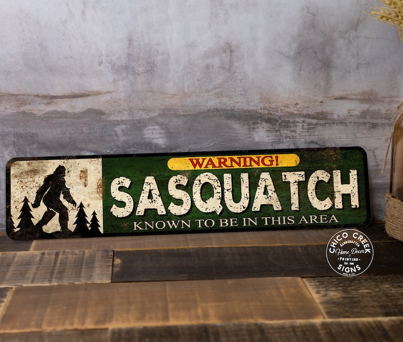 May include: A rectangular metal sign with a distressed finish. The sign features a silhouette of Bigfoot, the text "WARNING! SASQUATCH KNOWN TO BE IN THIS AREA," and a logo. The color scheme includes green, yellow, white, and brown.