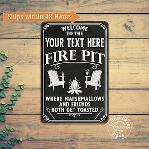 May include: Black and white metal sign with the text "Welcome to the Your Text Here Fire Pit" and an illustration of a campfire with two chairs. The text below the illustration reads "Where marshmallows and friends both get toasted".