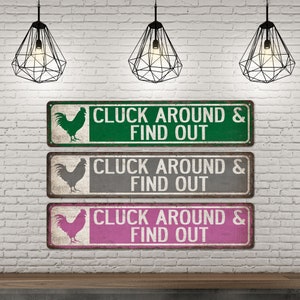 K&ouml;nnte beinhalten: Drei rustikale Metallschildchen mit einer Hahnensilhouette und dem Text "CLUCK AROUND & FIND OUT" in wei&szlig;en Buchstaben. Die Schilder sind gr&uuml;n, grau und rosa.