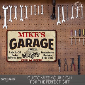 May include: A vintage-style metal sign with the text "MIKE'S GARAGE" in red and black lettering. The sign features a cartoon image of a classic car and lists services offered, including "Lube & Oil", "Brakes", "Tubes & Tires", "Transmission", "Radiator", and "Body Work". The sign also states "OPEN 24 HOURS".