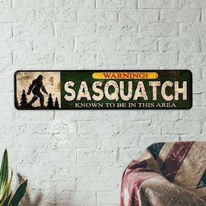 May include: A rectangular metal sign with a distressed finish. The sign features a silhouette of Bigfoot with trees, and the words "WARNING! SASQUATCH KNOWN TO BE IN THIS AREA" in white lettering on a green background.