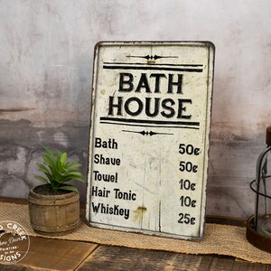 May include: A vintage-style metal sign with a white background and black lettering that reads "BATH HOUSE". The sign lists prices for various services, including bath, shave, towel, hair tonic, and whiskey.