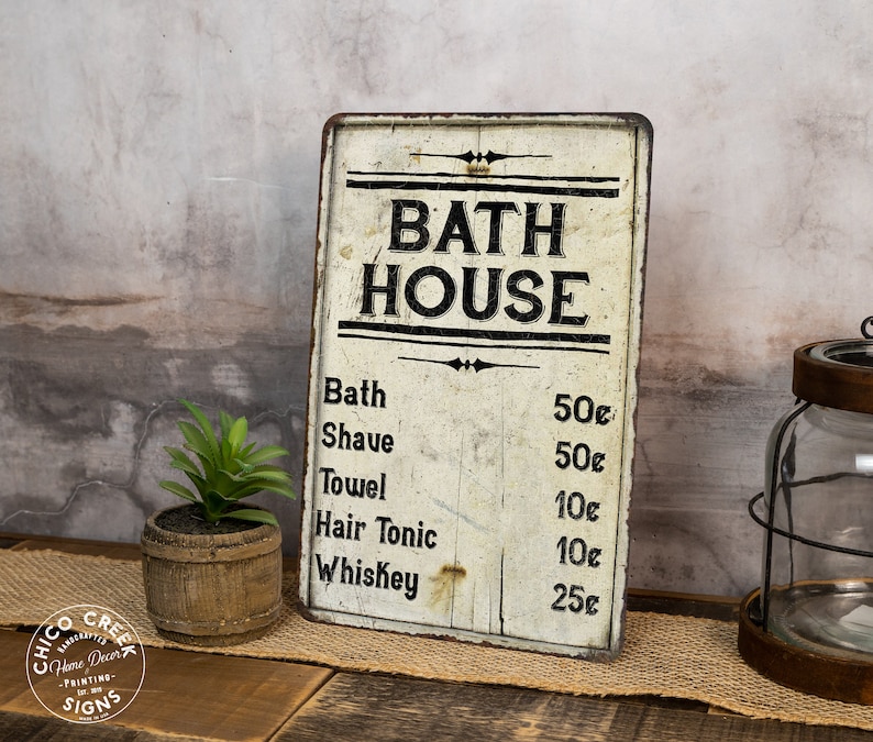May include: A vintage-style metal sign with a white background and black lettering that reads "BATH HOUSE". The sign lists prices for various services, including bath, shave, towel, hair tonic, and whiskey.