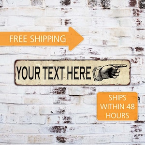 May include: A rectangular metal sign with the text "YOUR TEXT HERE" in black, featuring a pointing hand illustration. The sign has a distressed, vintage look, set against a white brick wall.
