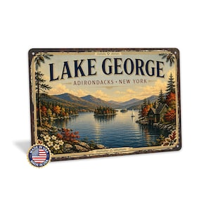 May include: A rectangular metal sign featuring a landscape illustration of Lake George in the Adirondacks, New York. The scene depicts a calm lake, mountains, trees, and a small building. The text reads LAKE GEORGE ADIRONDACKS NEW YORK.
