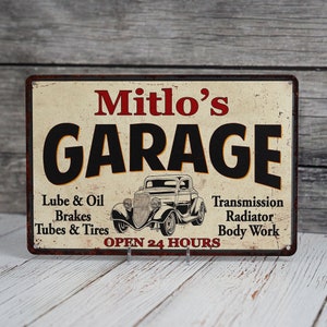 May include: A vintage-style metal sign with a rusty finish, advertising "Mitlo's Garage". The sign features a black and white illustration of a classic car, with the text "Open 24 Hours" below. The sign lists services offered, including "Lube & Oil", "Brakes", "Tubes & Tires", "Transmission", "Radiator", and "Body Work".