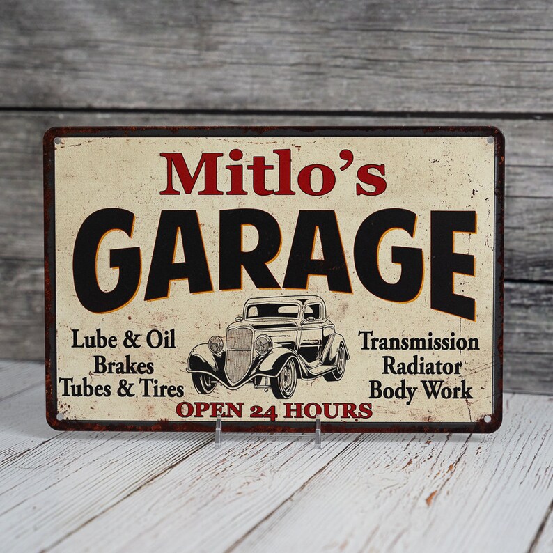 May include: A vintage-style metal sign with a rusty finish, advertising "Mitlo's Garage". The sign features a black and white illustration of a classic car, with the text "Open 24 Hours" below. The sign lists services offered, including "Lube & Oil", "Brakes", "Tubes & Tires", "Transmission", "Radiator", and "Body Work".